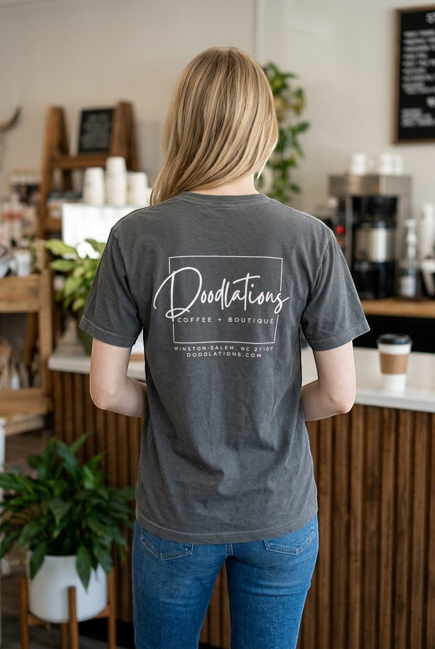 Woman wearing a charcoal grey Doodlations Coffee Bar & Boutique graphic tee with white logo on the back in a cafe