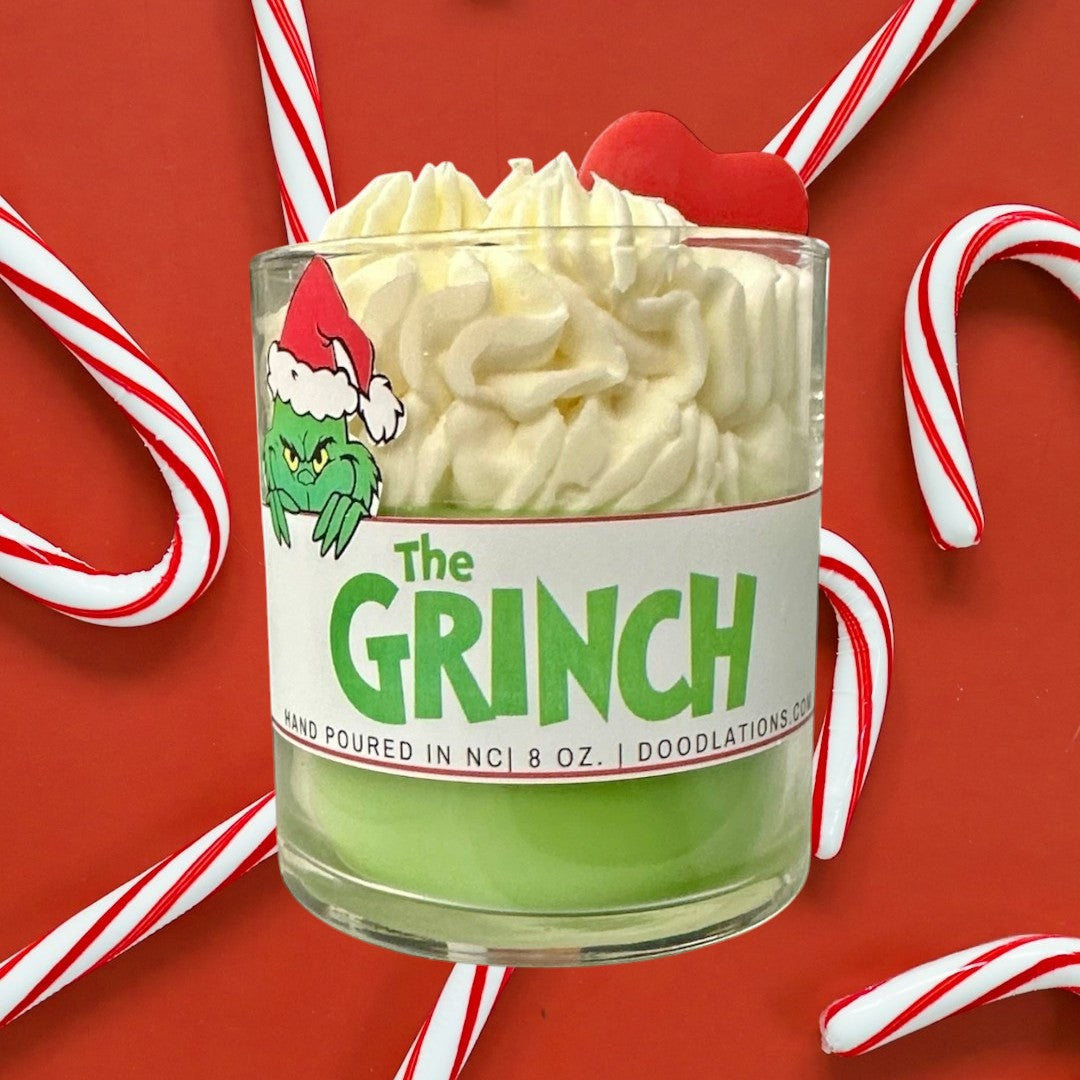 Doodlations Coffee Bar & Boutique The Grinch candle with green wax and whipped cream top, decorated with a Grinch sticker and red heart.