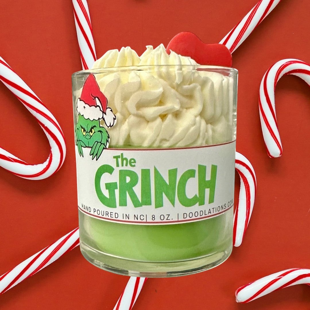 Doodlations Coffee Bar & Boutique The Grinch candle with green wax and whipped cream top, decorated with a Grinch sticker and red heart.