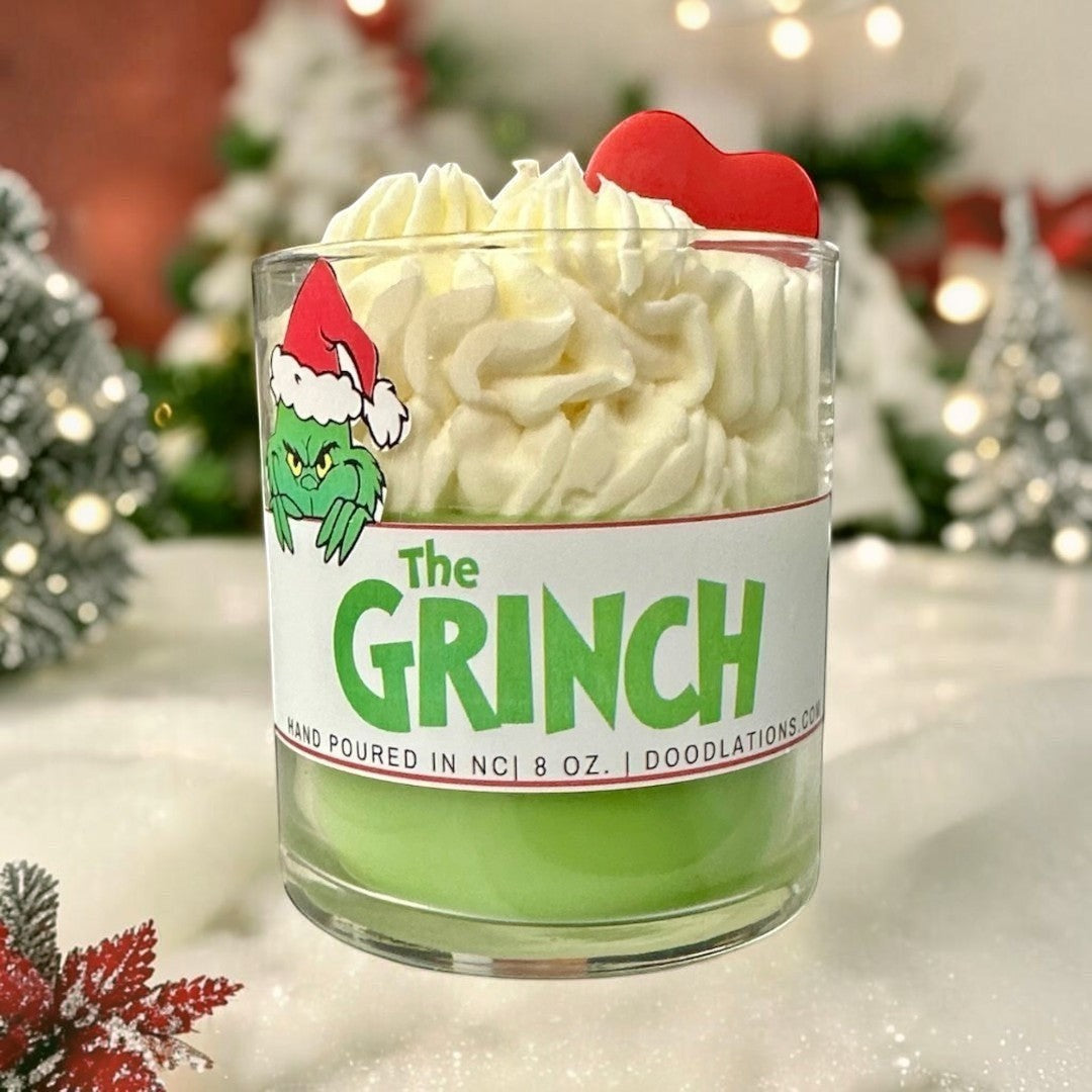 Doodlations Coffee Bar & Boutique The Grinch candle with green wax and white whipped wax topping, decorated with a Grinch sticker and red heart.