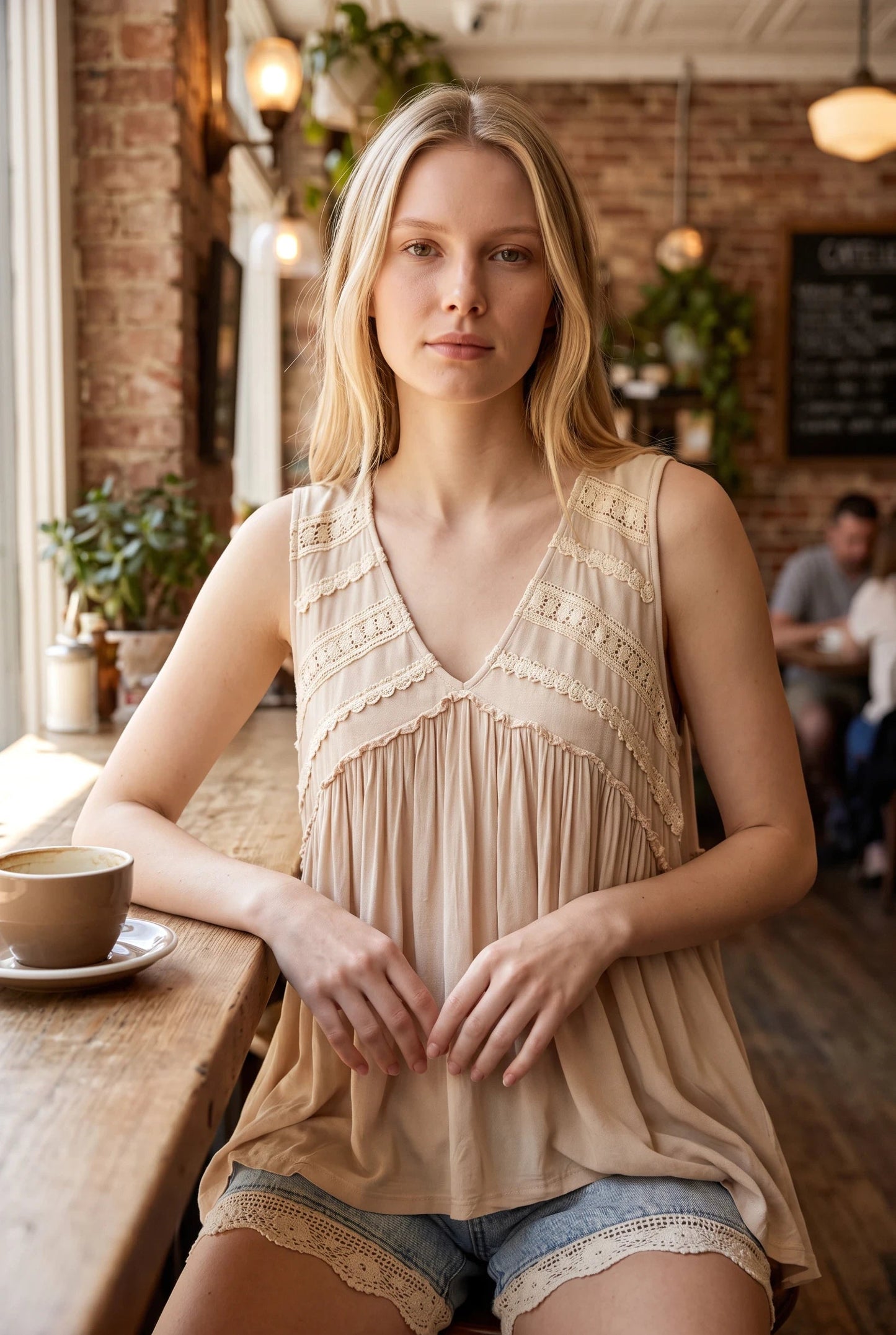 A blonde woman wearing a Doodlations Coffee Bar & Boutique ivory sleeveless V-neck tank top with lace trim in a cafe.