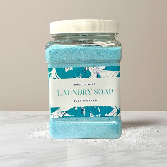 Doodlations Coffee Bar & Boutique Just Washed powdered laundry soap in a clear plastic jar with a white lid. Blue powder fills the jar.