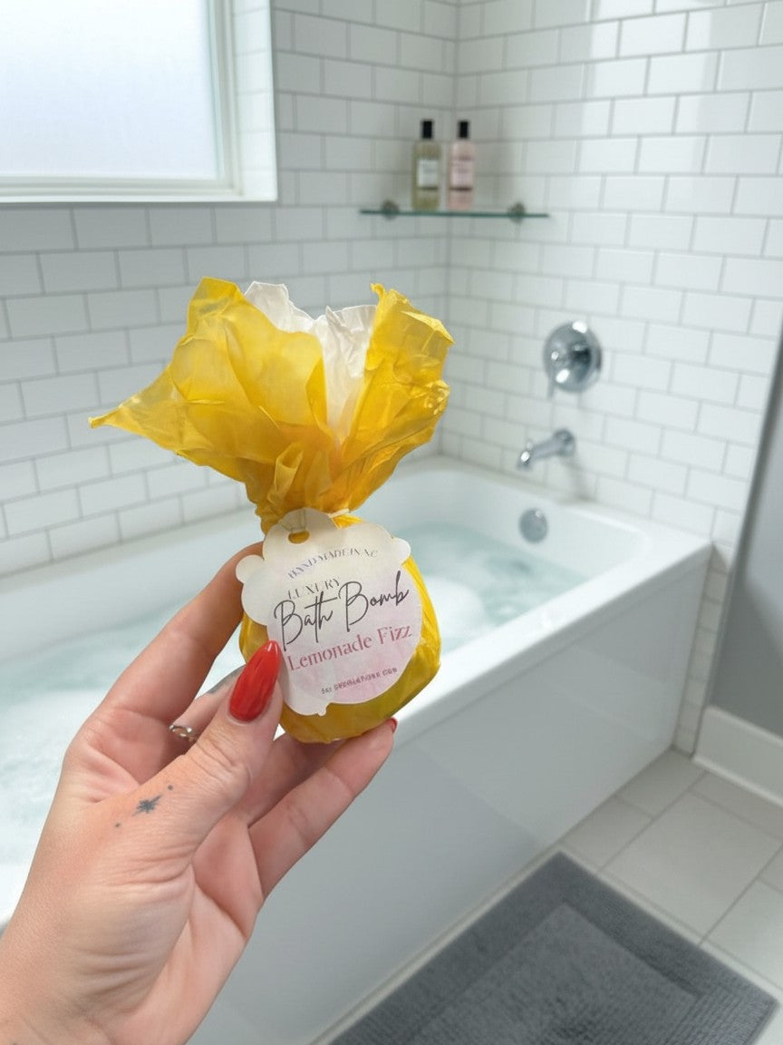 Hand holding a yellow Doodlations Coffee Bar & Boutique Lemonade Fizz bath bomb in a bright white tiled bathroom.