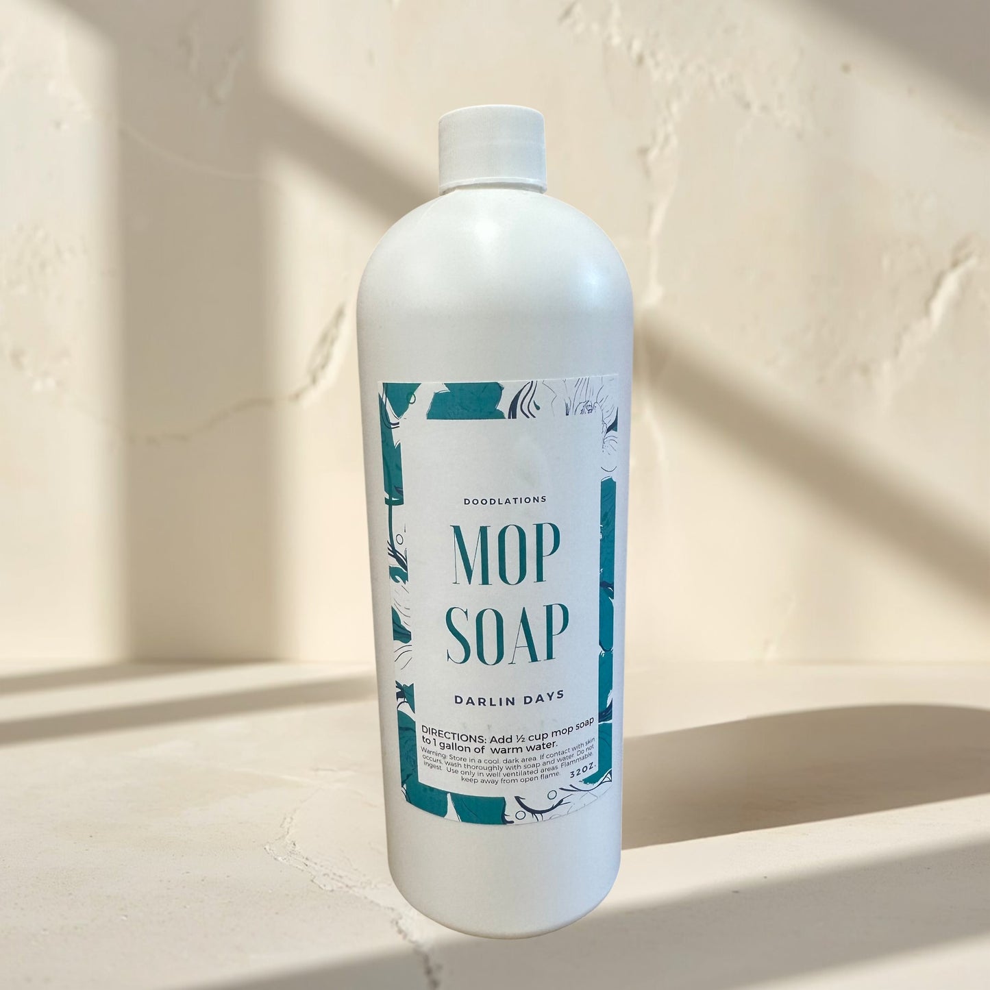 Doodlations Coffee Bar & Boutique Darlin’ Days concentrated mop soap in a white bottle with a teal and white label. A gold mop is behind it.