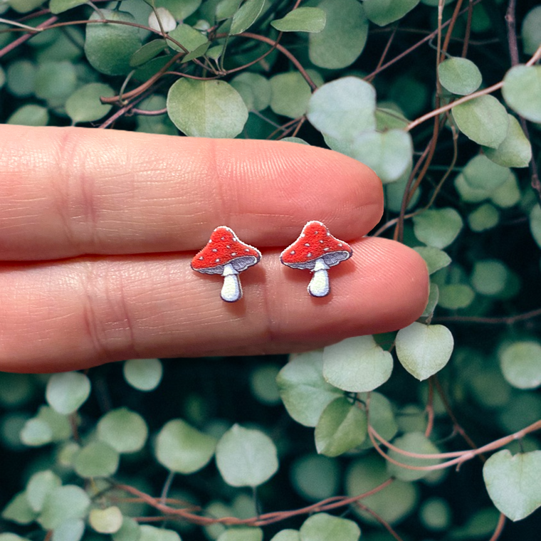 Doodlations Coffee Bar & Boutique handmade red and white mushroom stud earrings held against a green leafy background.