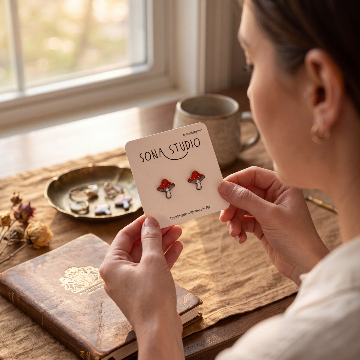 Doodlations Coffee Bar & Boutique handmade red and white mushroom stud earrings on a display card held by a woman