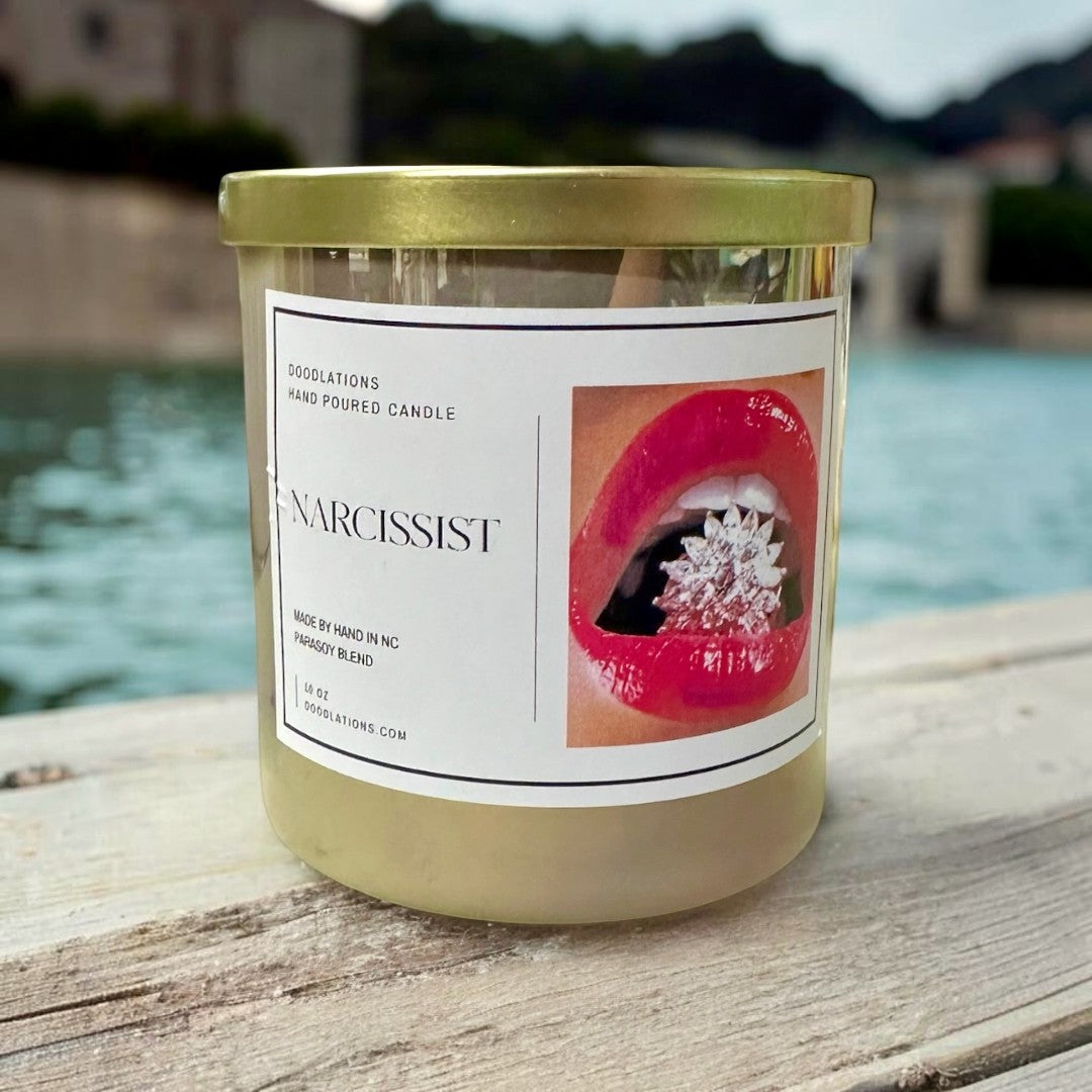 Doodlations Coffee Bar & Boutique Narcissist hand-poured soy blend candle in a clear jar with a gold lid, featuring a label with red lips and a sparkling jewel.