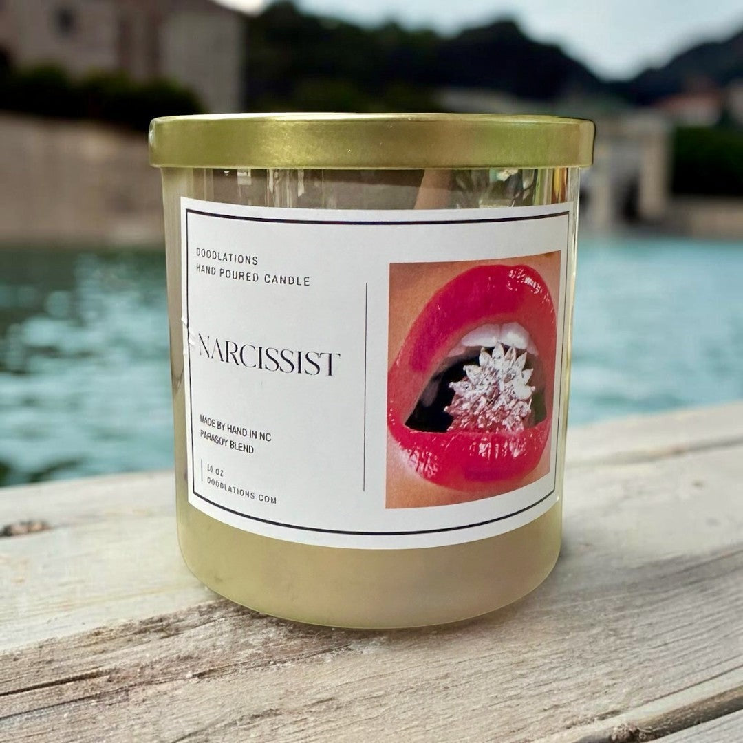 Doodlations Coffee Bar & Boutique Narcissist hand-poured soy blend candle in a clear jar with a gold lid, featuring a label with red lips and a sparkling jewel.
