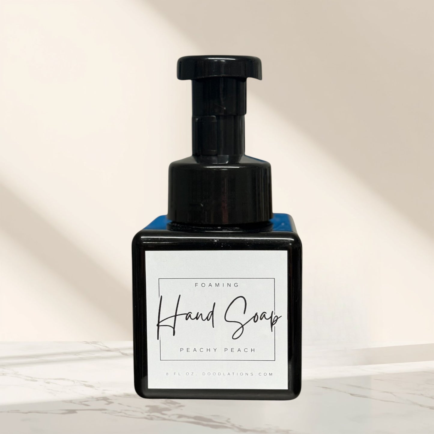 Doodlations Peachy Peach foaming hand soap in a black square bottle with a black pump dispenser. Surrounded by green artificial plants.