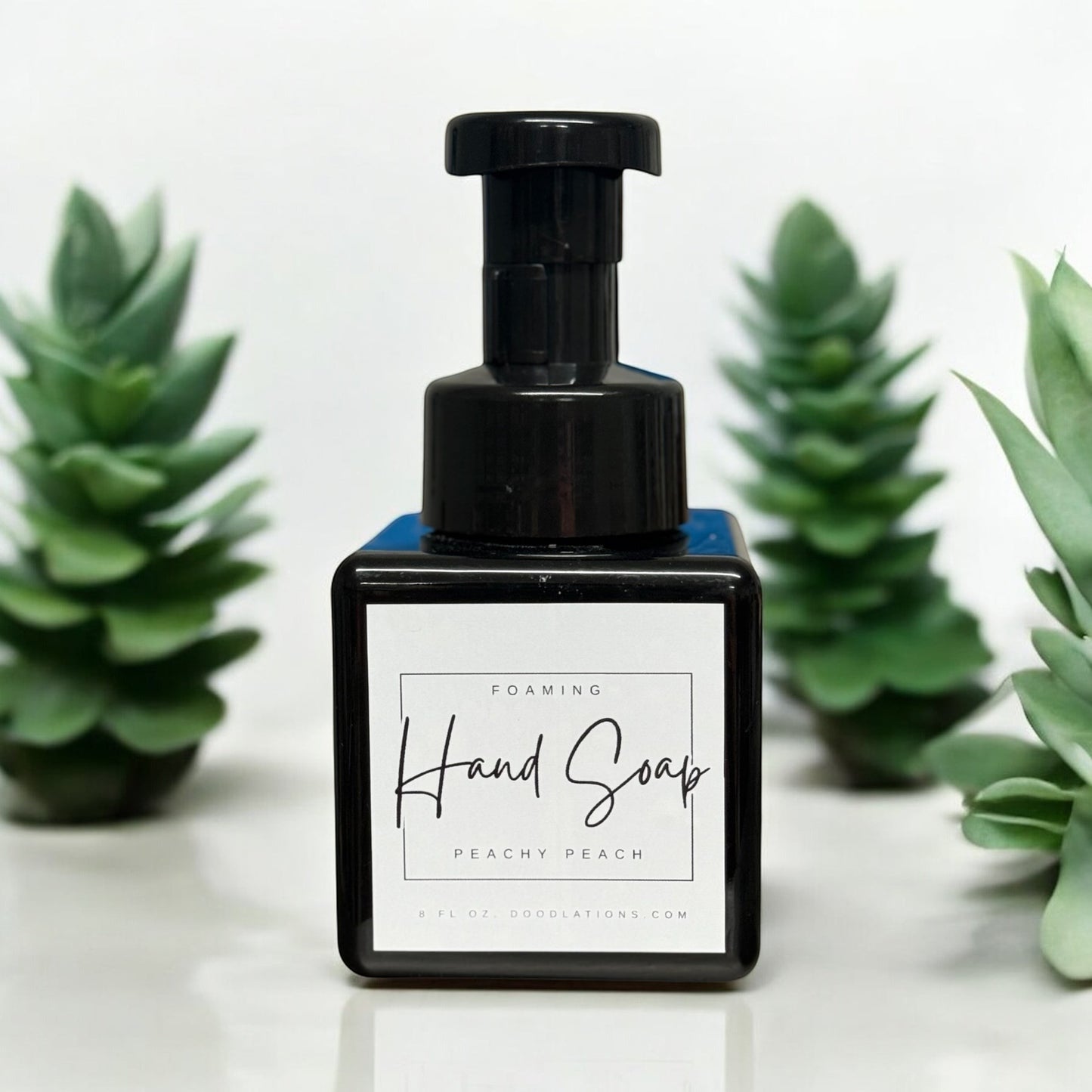 Doodlations Peachy Peach foaming hand soap in a black square bottle with a black pump dispenser. Surrounded by green artificial plants.