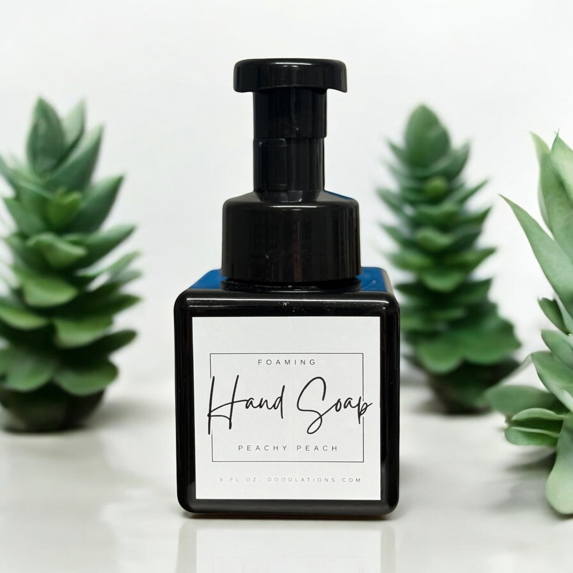 Doodlations Peachy Peach foaming hand soap in a black square bottle with a black pump dispenser. Surrounded by green artificial plants.