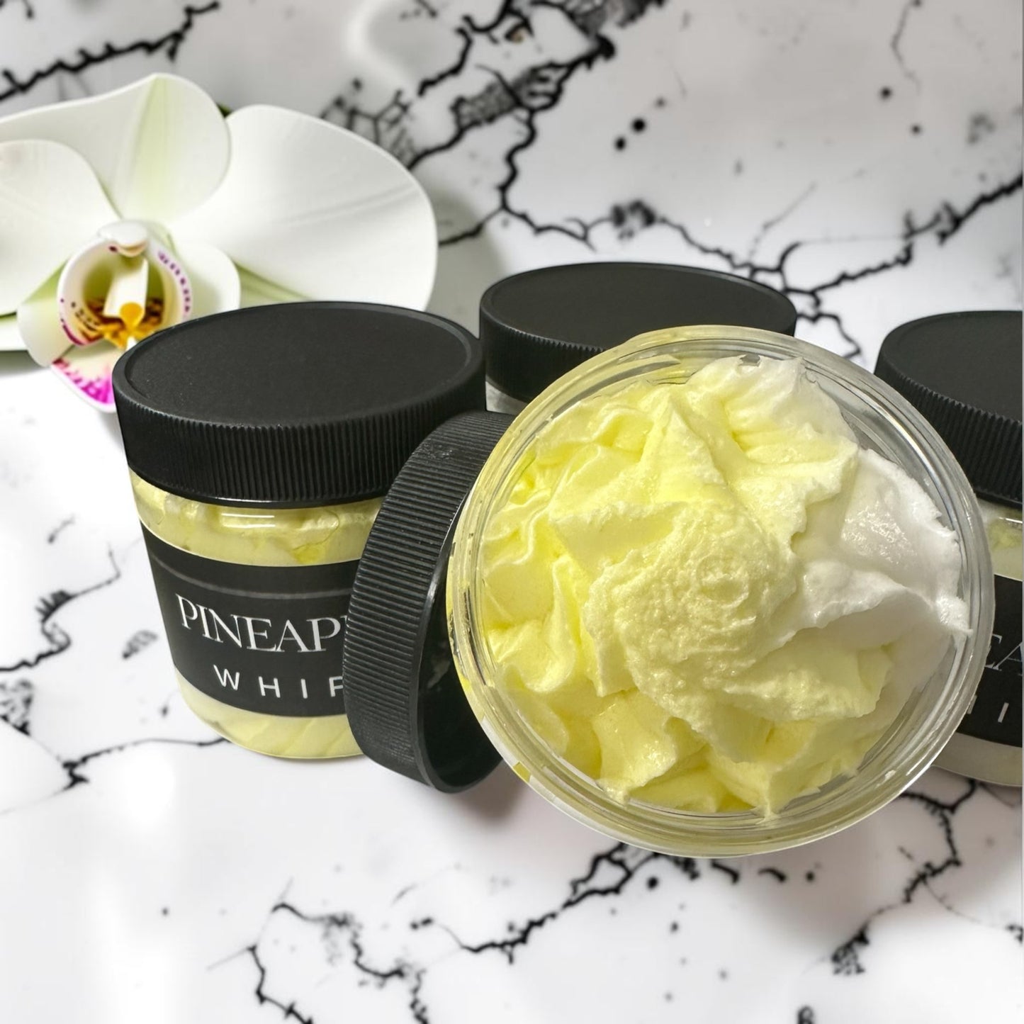Doodlations Coffee Bar & Boutique Pineapple Mango whipped soap in clear jars with black lids on a marble surface.