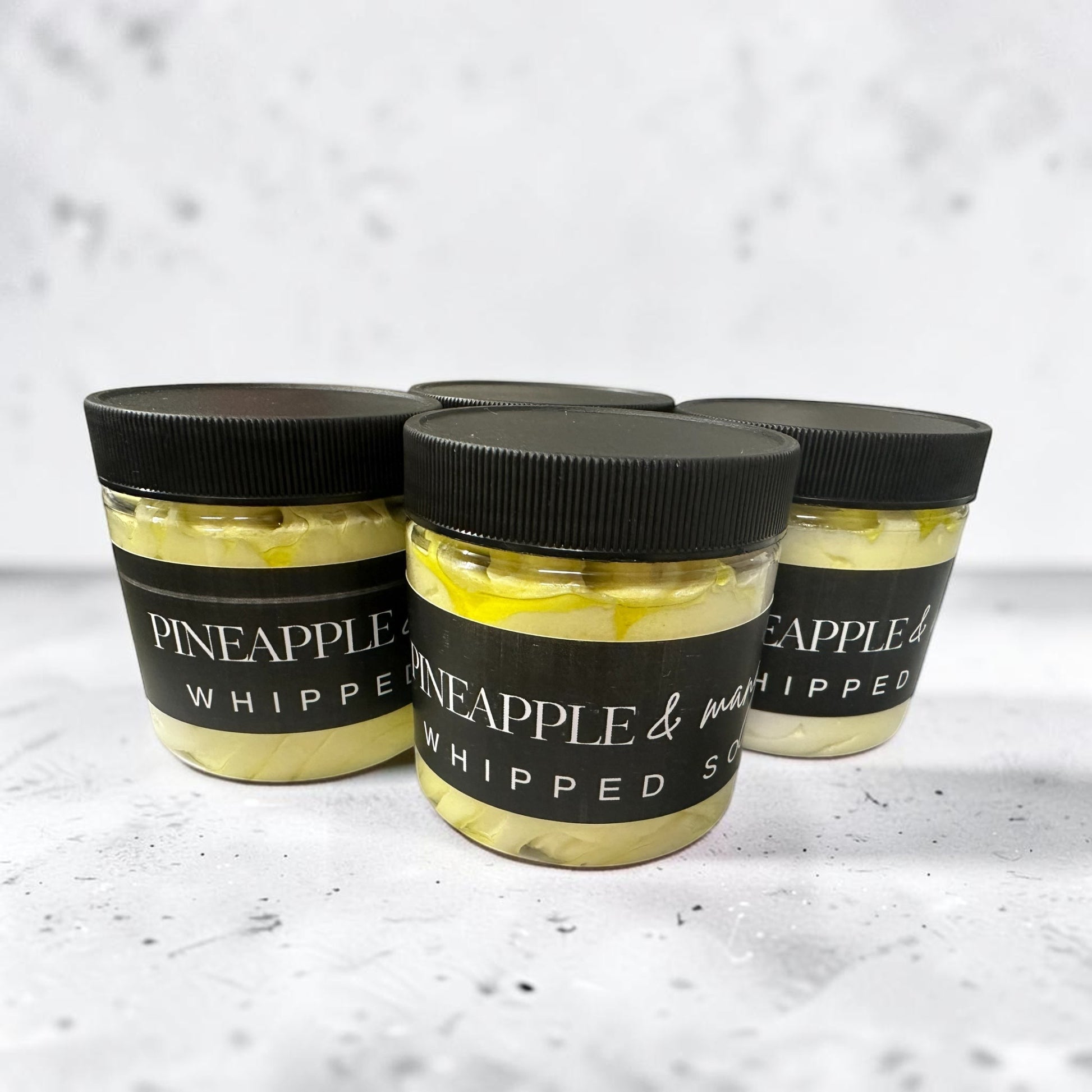 Doodlations Coffee Bar & Boutique Pineapple Mango whipped soap in clear jars with black lids on a textured surface.