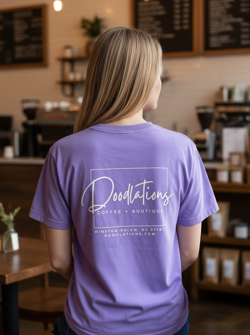 Woman wearing a purple Doodlations Coffee Bar & Boutique graphic tee with a white logo print on the back