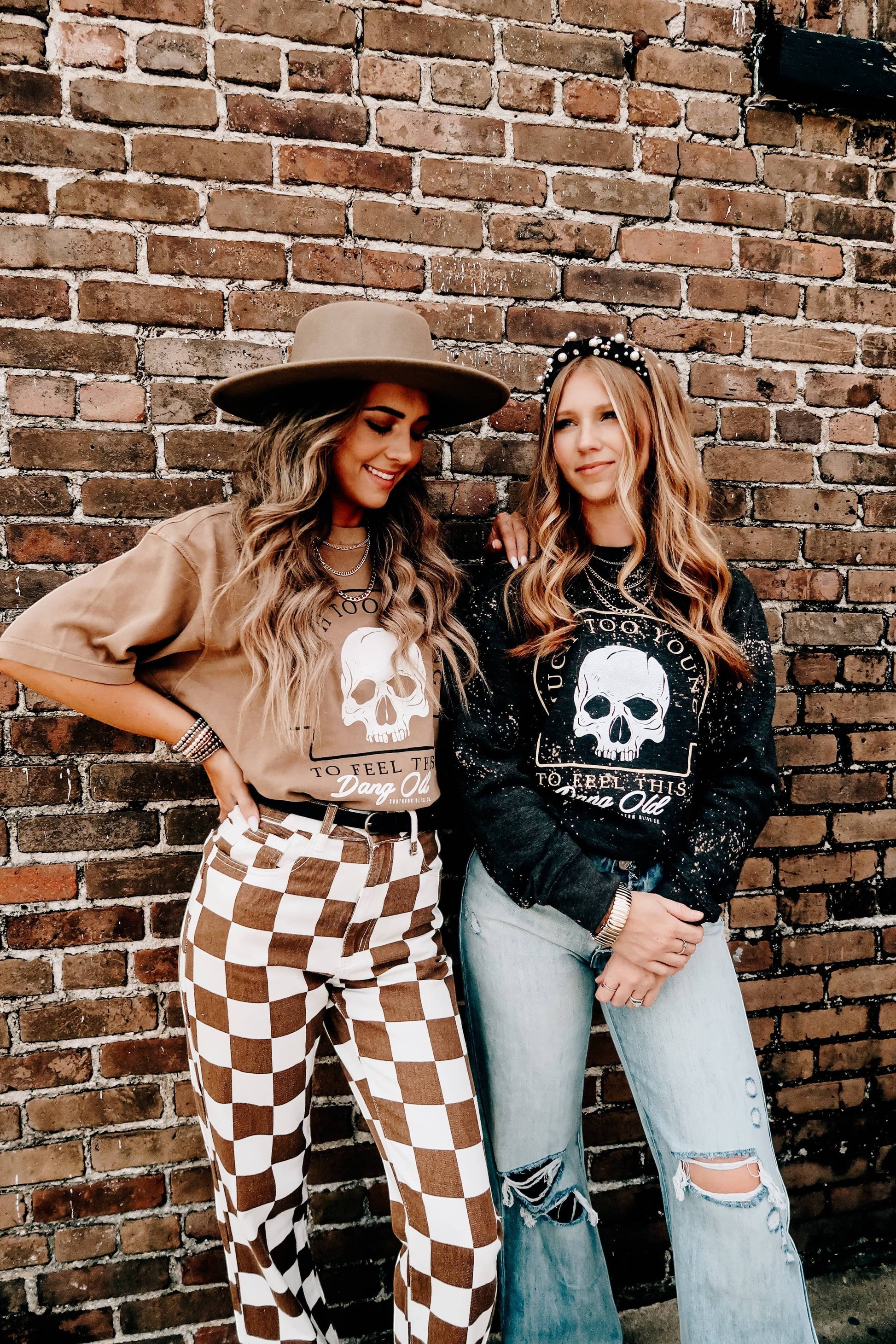 Doodlations Coffee Bar & Boutique graphic skull t-shirts in tan and black worn by two women against a brick wall