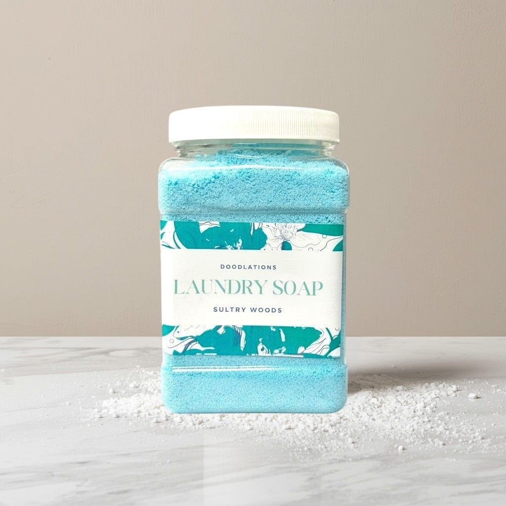 Doodlations Coffee Bar & Boutique Sultry Woods powder laundry soap in a clear plastic container with a white lid. Blue powder fills the container.