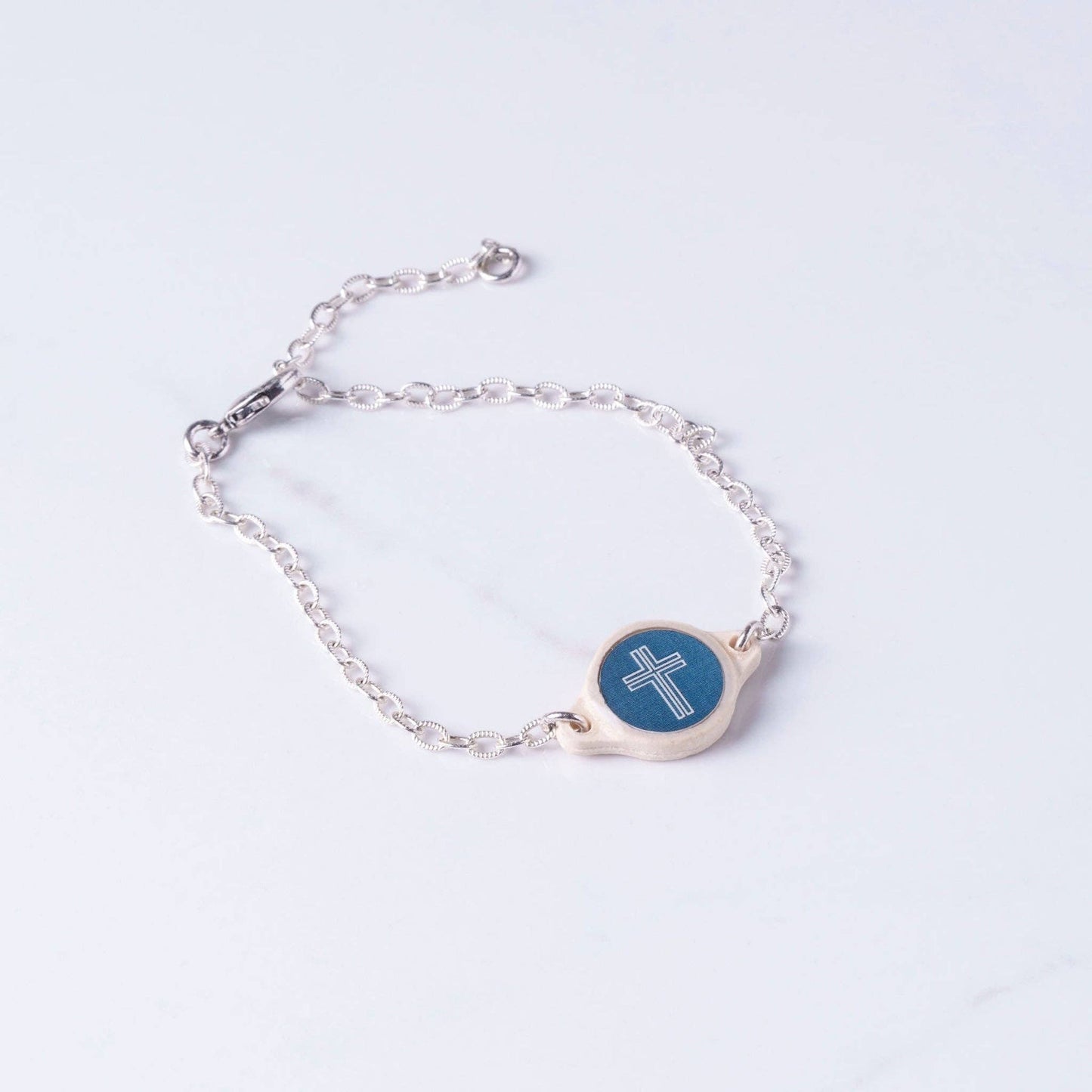 Doodlations Coffee Bar & Boutique Survivor Series bracelet with a silver chain and a cross charm on a moss green background.