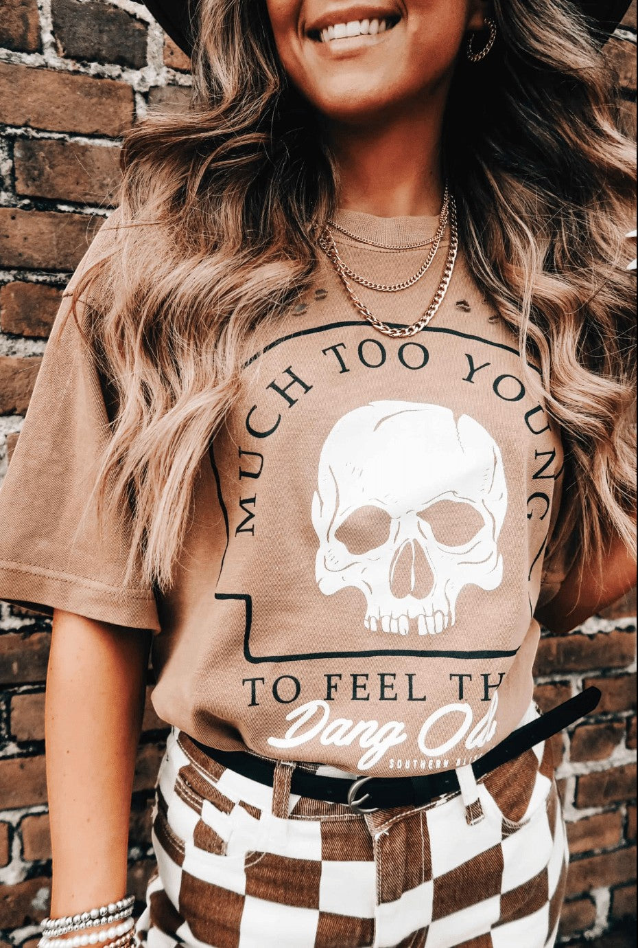 Doodlations Coffee Bar & Boutique tan graphic skull t-shirt with text on a woman wearing a black hat and gold chains