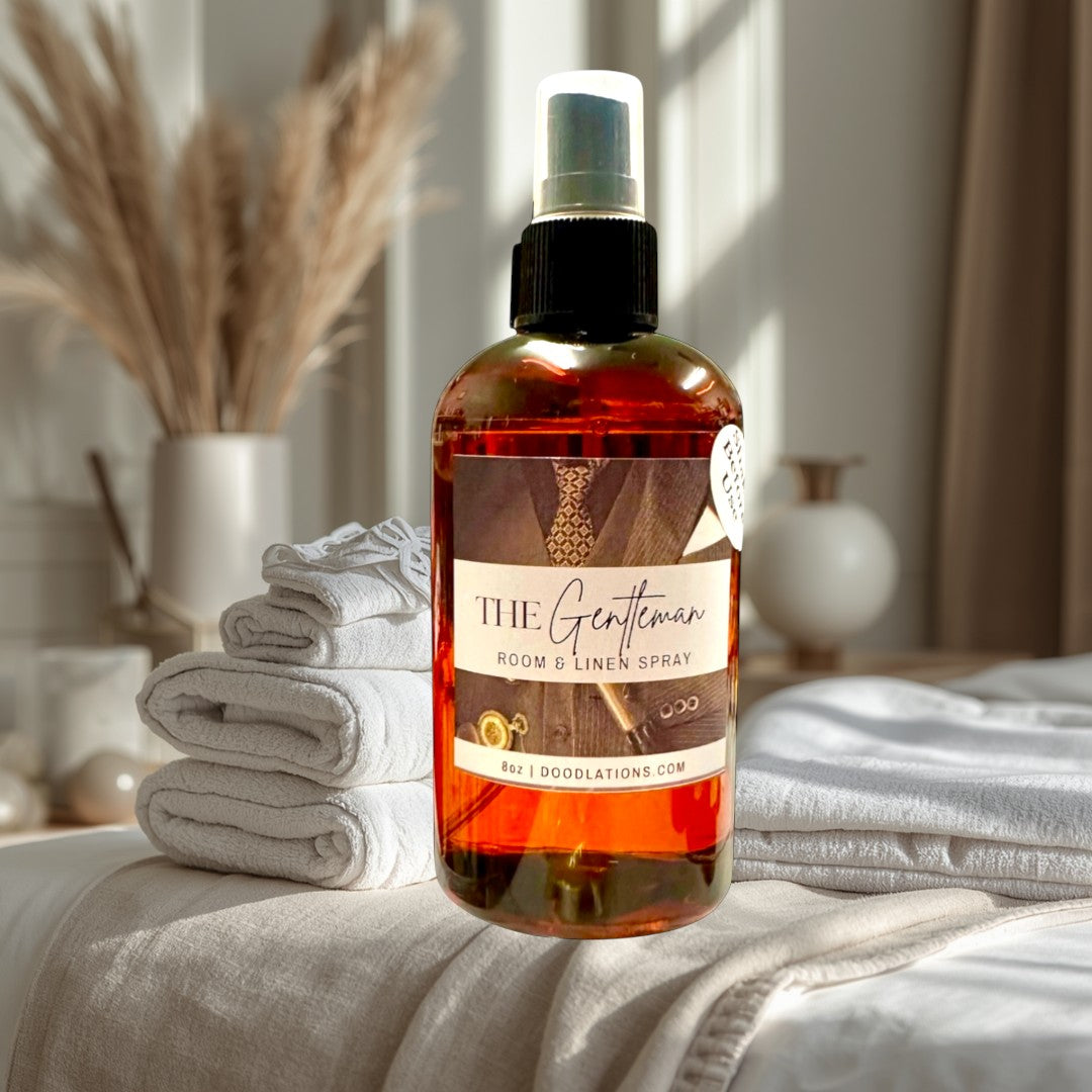Doodlations Coffee Bar & Boutique The Gentleman room and linen spray in amber glass bottle with black spray top. Scent notes: mahogany, blood orange, red pepper, bourbon vanilla, amber, tonka bean, sandalwood.