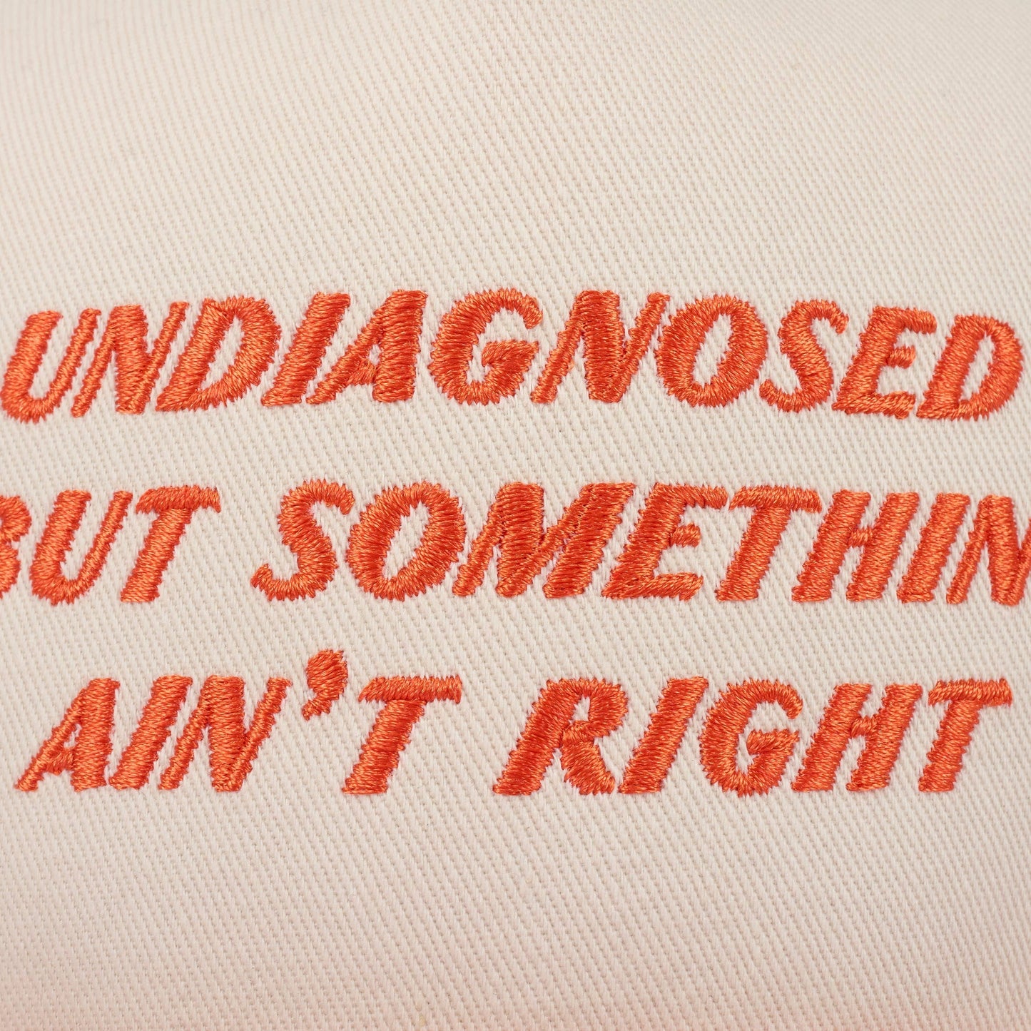 Doodlations Coffee Bar & Boutique cream trucker hat with orange embroidered text "UNDIAGNOSED BUT SOMETHIN AIN'T RIGHT"