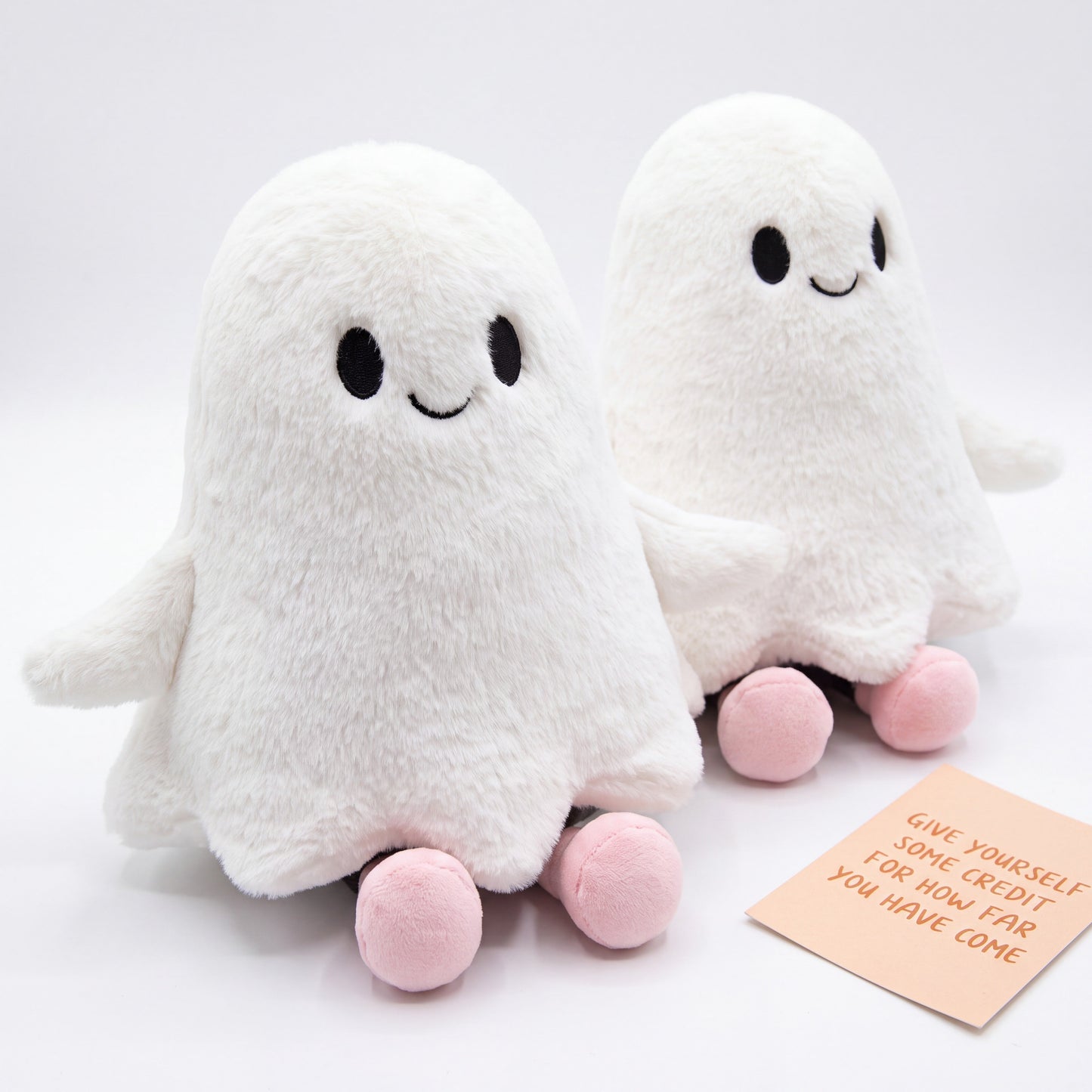 Two Doodlations Coffee Bar & Boutique white plush ghost toys with friendly embroidered faces and small pink feet.