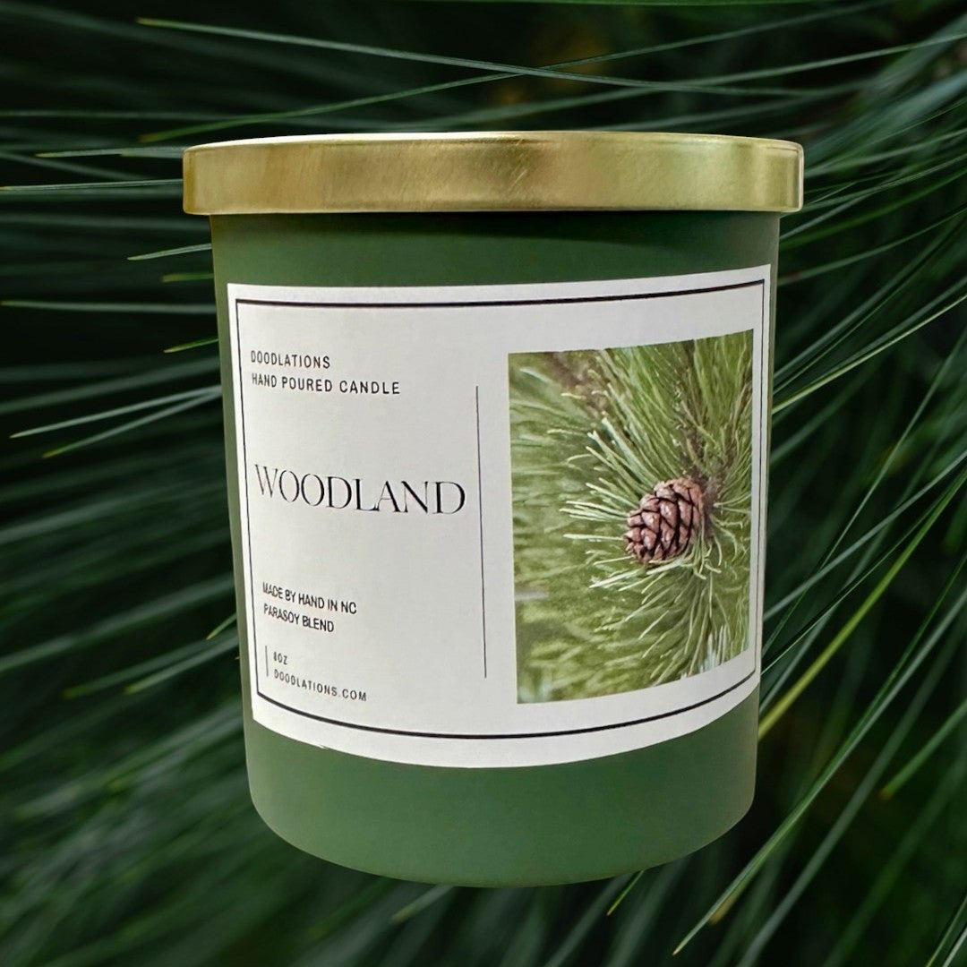 Doodlations Coffee Bar & Boutique Woodland hand-poured para-soy blend candle with pine cone imagery. Green jar, gold lid.