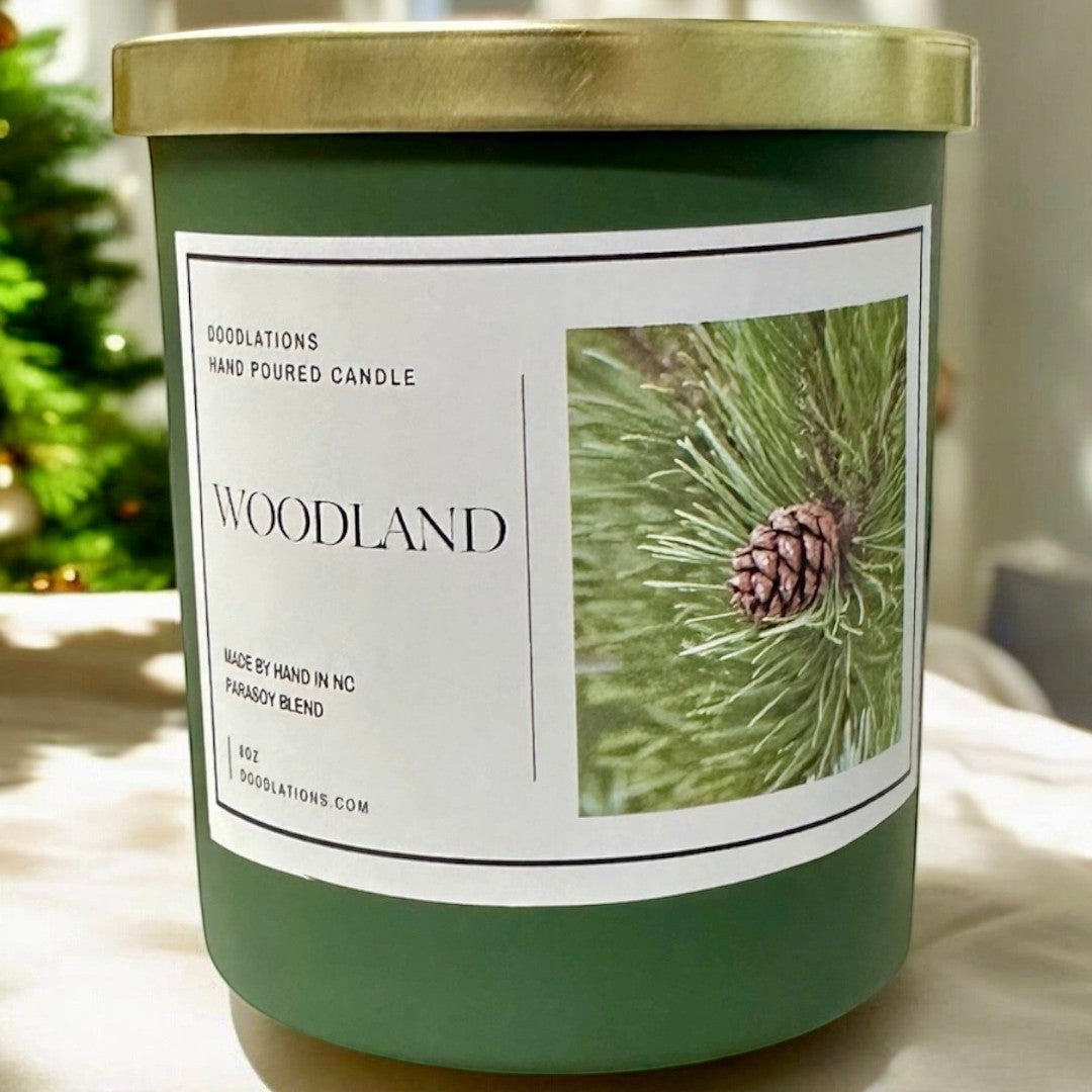 Doodlations Coffee Bar & Boutique Woodland candle in a green jar with a gold lid, featuring a pine cone image.