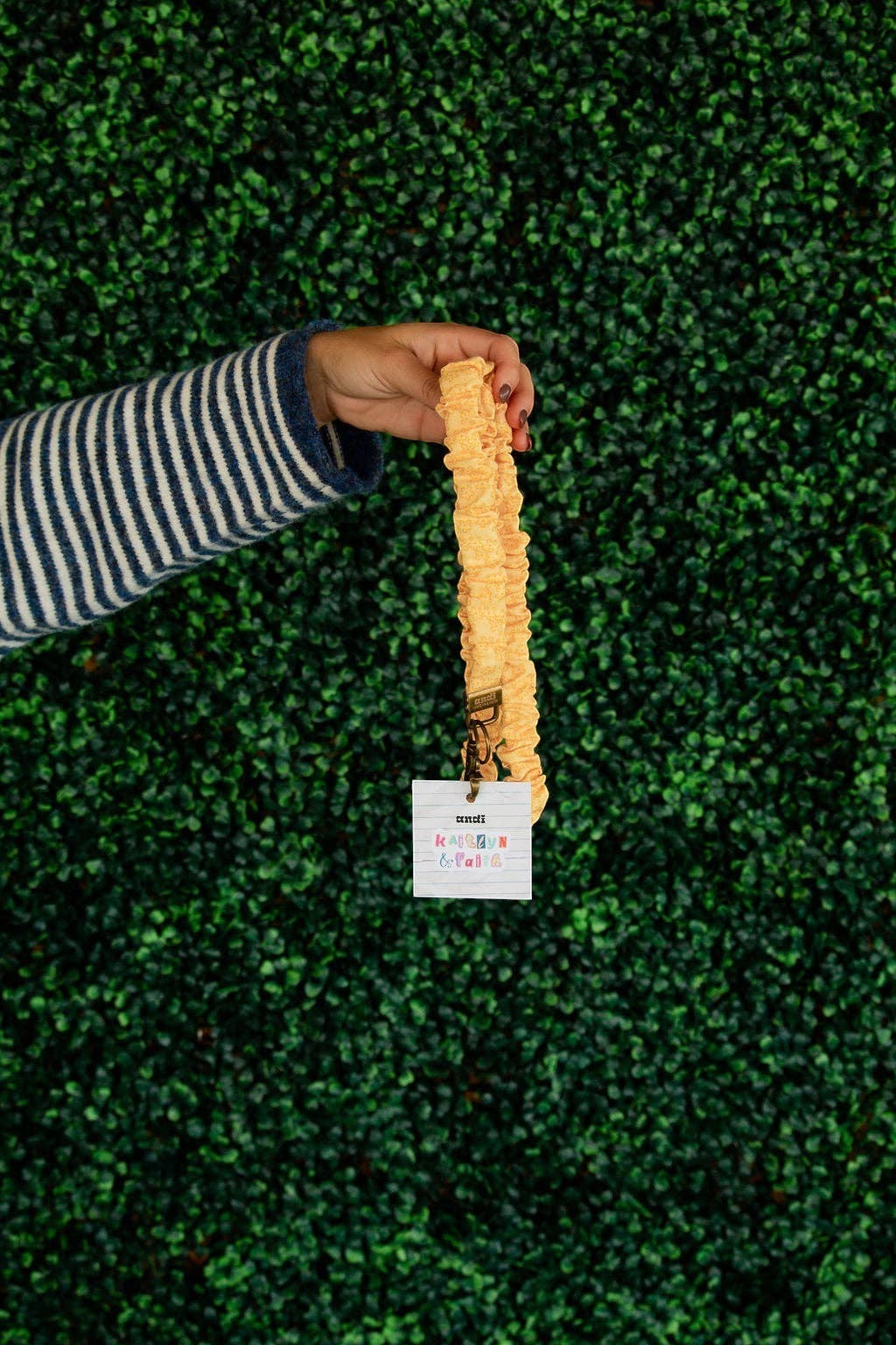 Doodlations Coffee Bar & Boutique yellow floral linen lanyard with a gold clasp held against a green hedge background