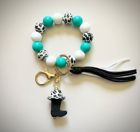 Doodlations Coffee Bar & Boutique cow print boot charm keychain bracelet with turquoise, white, and cow-print beads, and a black boot charm with tassel.