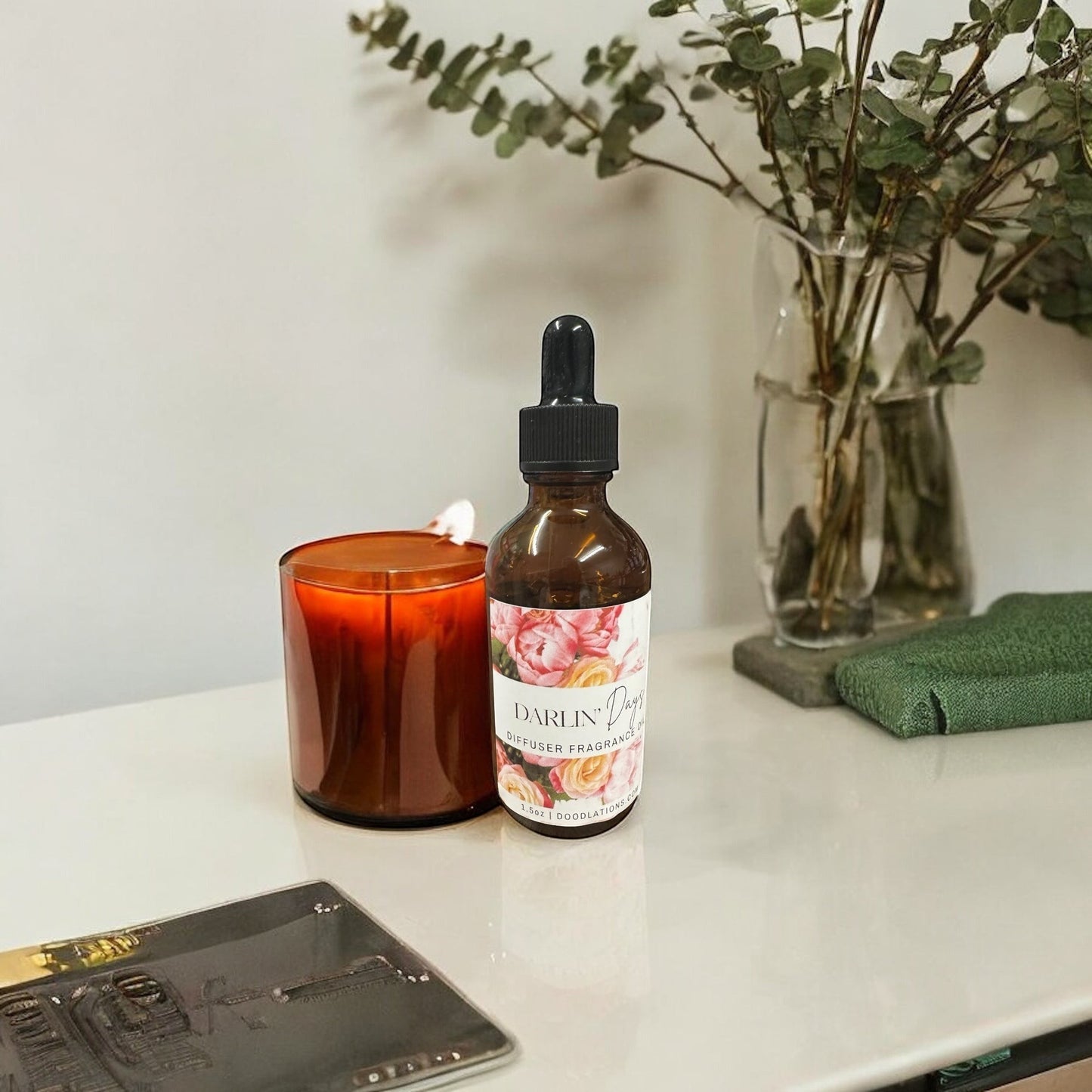 Doodlations amber glass bottle with dropper of floral diffuser fragrance oil next to a lit amber candle.