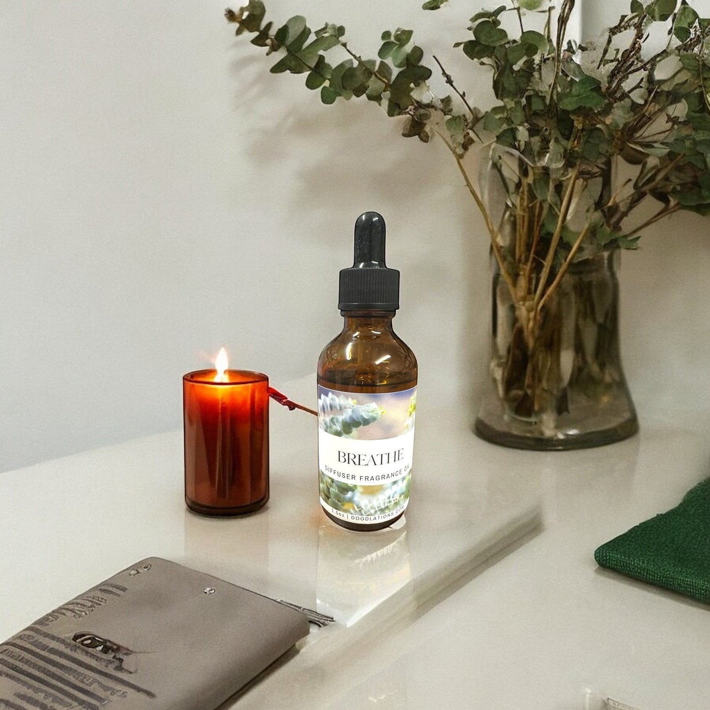 Doodlations Coffee Bar & Boutique Breathe diffuser fragrance oil in amber glass bottle with dropper, next to a lit candle and eucalyptus branches.