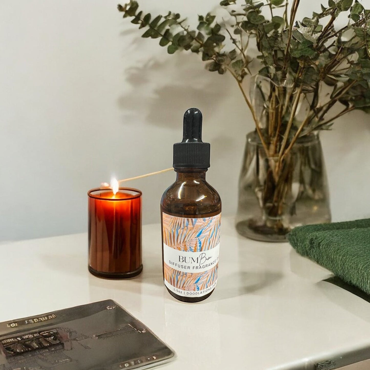 Doodlations Coffee Bar & Boutique amber glass bottle of diffuser fragrance oil with dropper, next to a lit candle and eucalyptus in a vase.