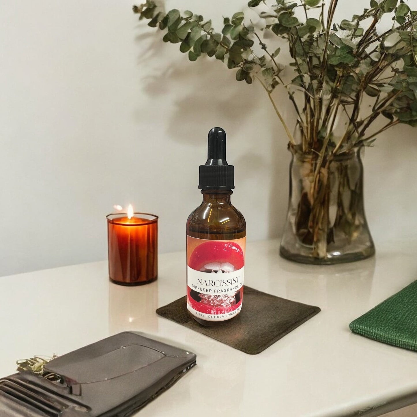 Doodlations Coffee Bar & Boutique Narcissist fragrance oil bottle with dropper, amber glass, red lips label, next to lit candle and eucalyptus.