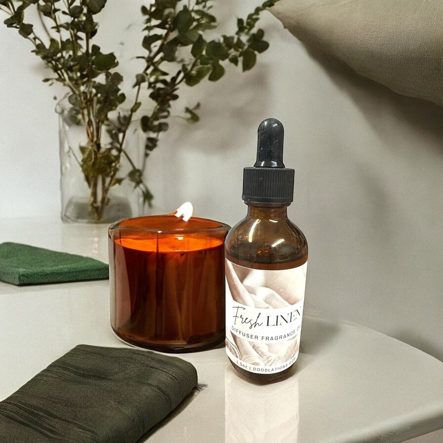 Doodlations Coffee Bar & Boutique Fresh Linen diffuser fragrance oil in amber dropper bottle next to a lit candle.