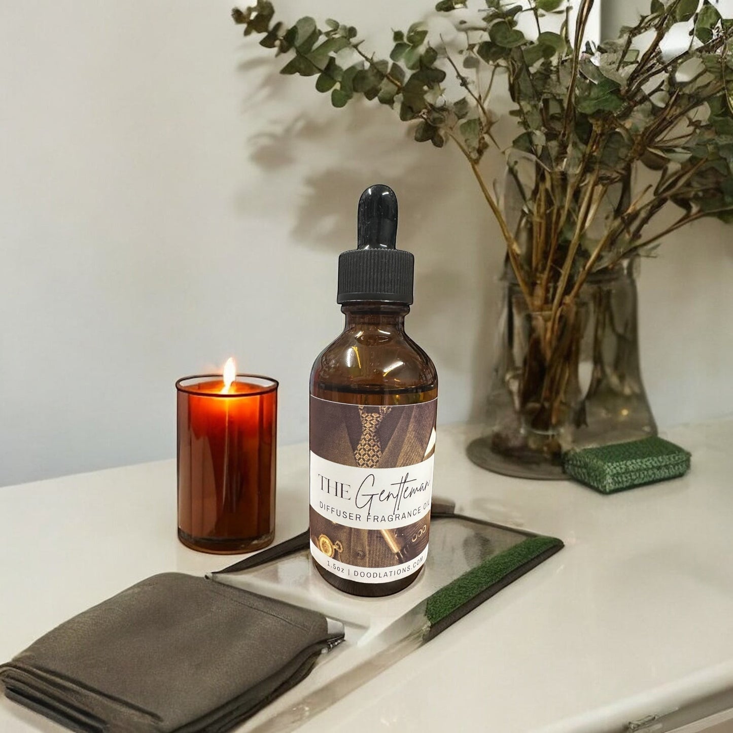 Doodlations Coffee Bar & Boutique The Gentleman diffuser fragrance oil in amber glass bottle with dropper. Includes lit candle and eucalyptus branches.