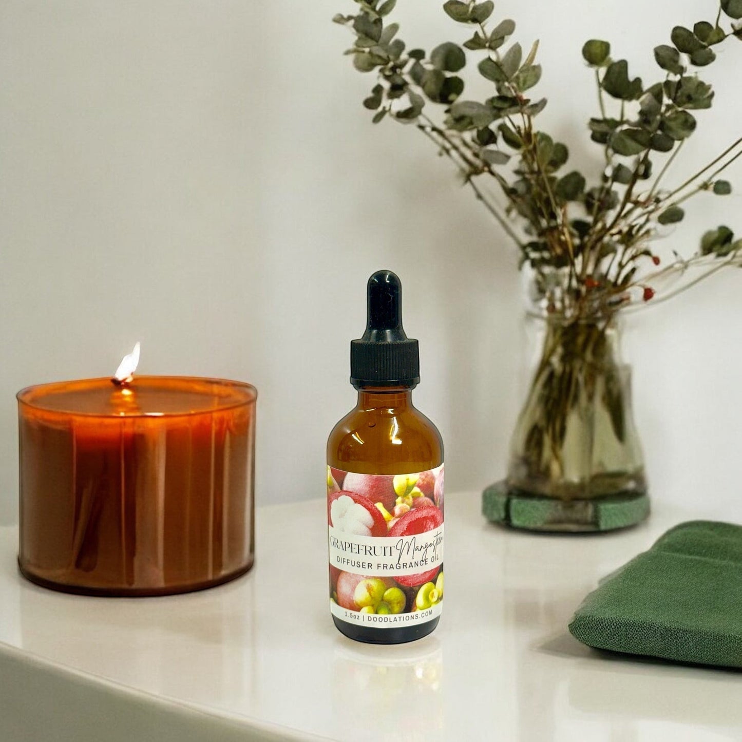 Doodlations Coffee Bar & Boutique Grapefruit Mangosteen diffuser fragrance oil bottle with dropper, next to a lit candle and eucalyptus.