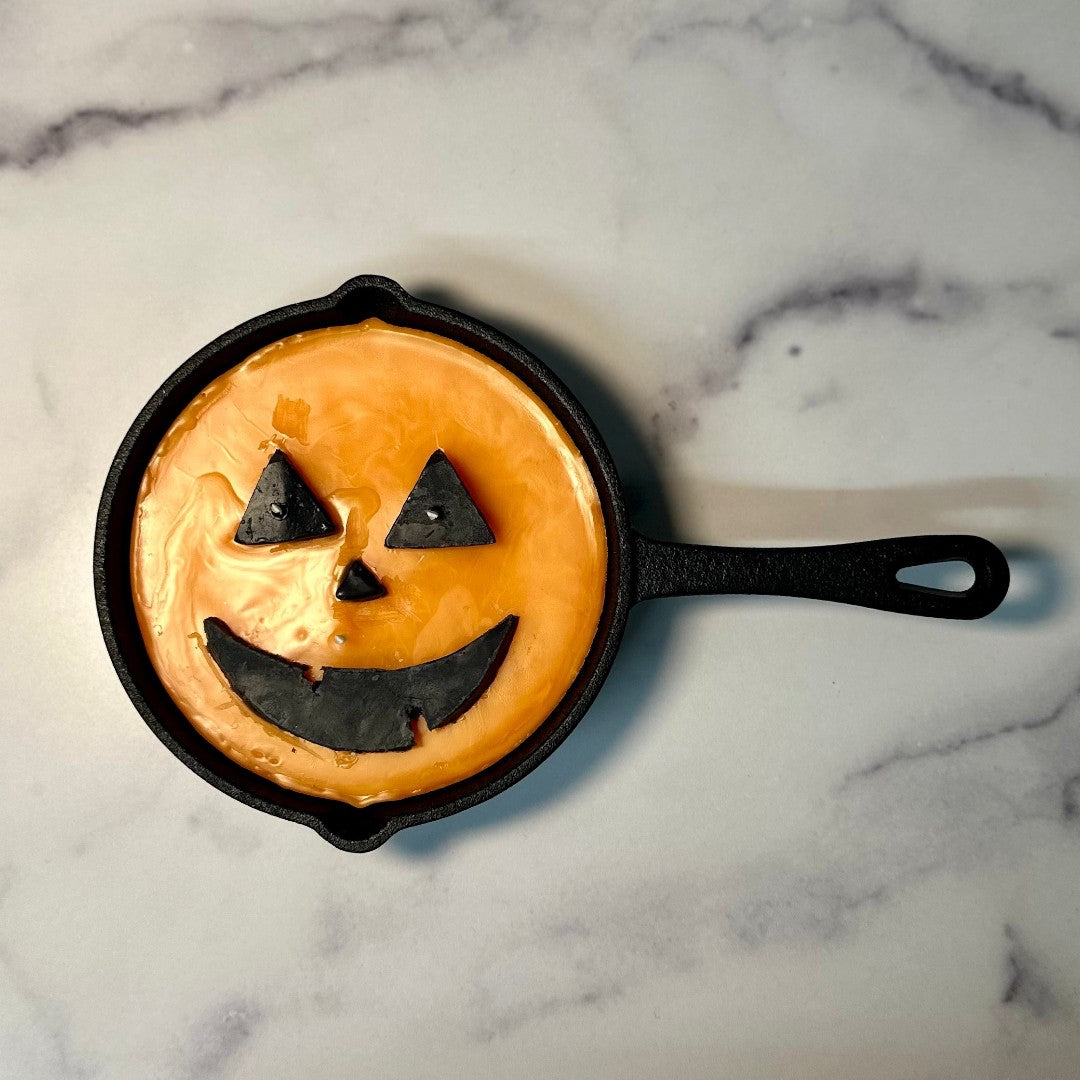 Doodlations unique candle in a cast iron skillet, shaped like a jack o' lantern with orange wax and black facial features.