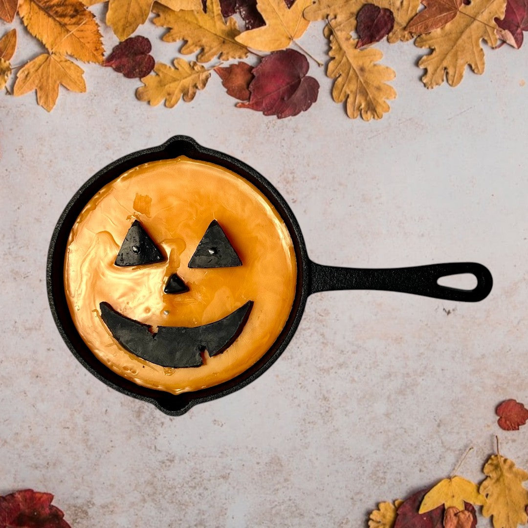 Doodlations unique candle in a cast iron skillet, shaped like a jack o’ lantern with orange wax and black facial features.