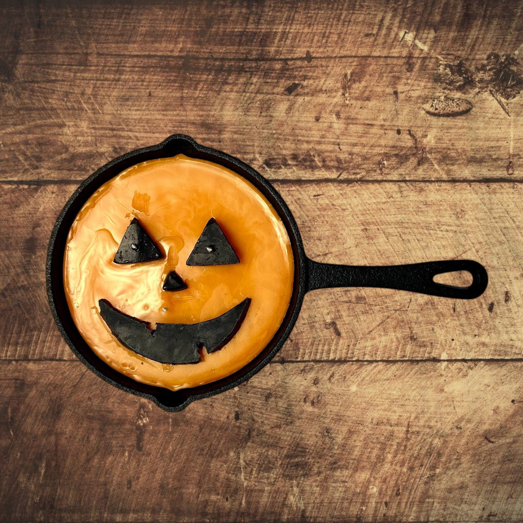 Doodlations unique candle in a cast iron skillet, shaped like a jack o’ lantern with orange wax and black facial features.