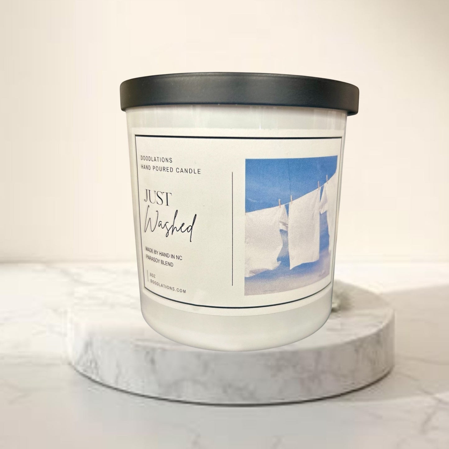 Doodlations Just Washed luxury boutique candle in a white jar with a black lid, featuring a label with white laundry hanging on a clothesline against a blue sky.