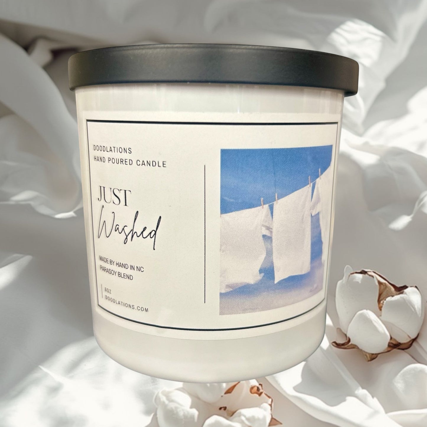 Doodlations Just Washed candle with a clean laundry scent. White jar with black lid and label featuring white clothes on a clothesline.