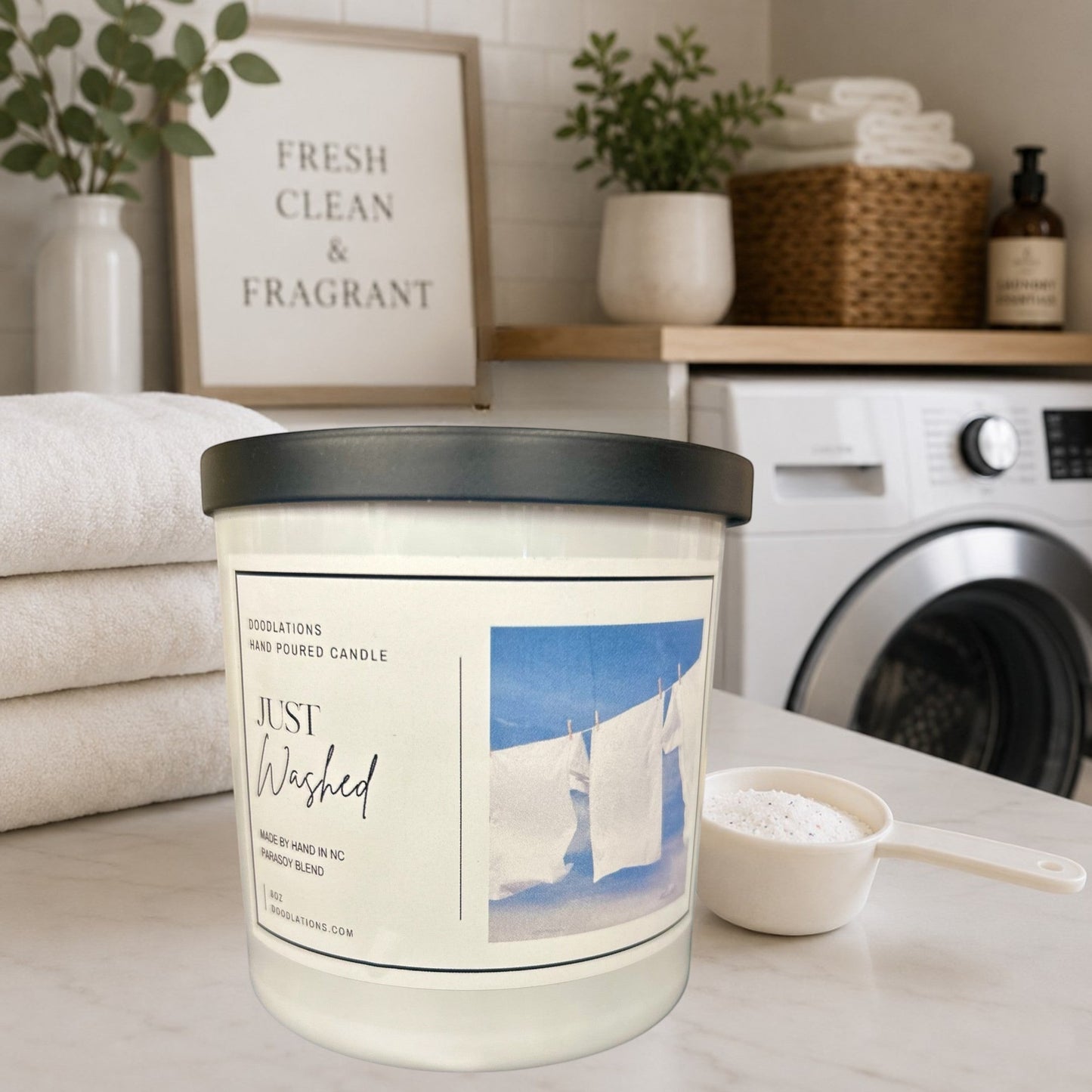 Doodlations Just Washed luxury boutique candle in a white jar with a black lid, featuring a label with white laundry hanging on a clothesline against a blue sky.