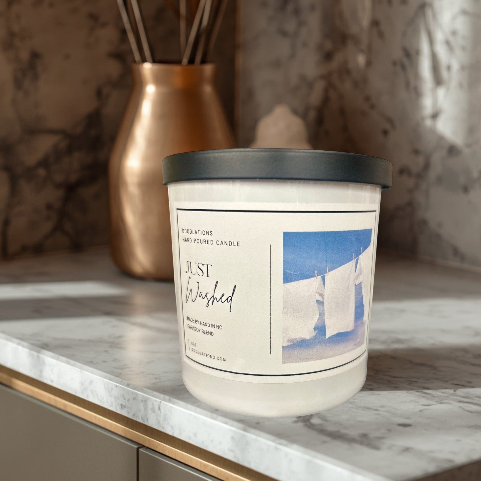 Doodlations Just Washed hand-poured parasoy blend candle with a fresh laundry scent. White candle in a clear jar with a black lid.