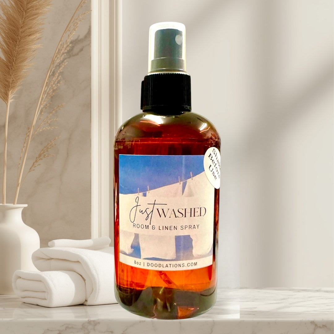 Doodlations Just Washed room and linen spray in amber bottle with black spray top. Scent notes: Mountain Air, Cotton Blossoms, Pink Jasmine, Sandalwood.