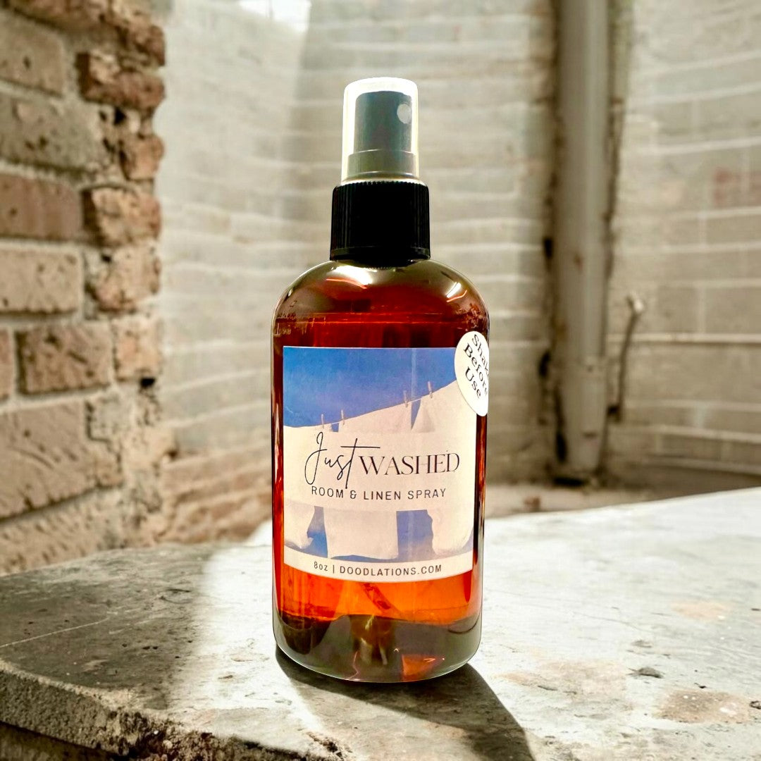 Doodlations Just Washed room and linen spray in amber bottle with label depicting clothes on a line. Scent notes: Mountain Air, Cotton Blossoms, Pink Jasmine, Sandalwood.
