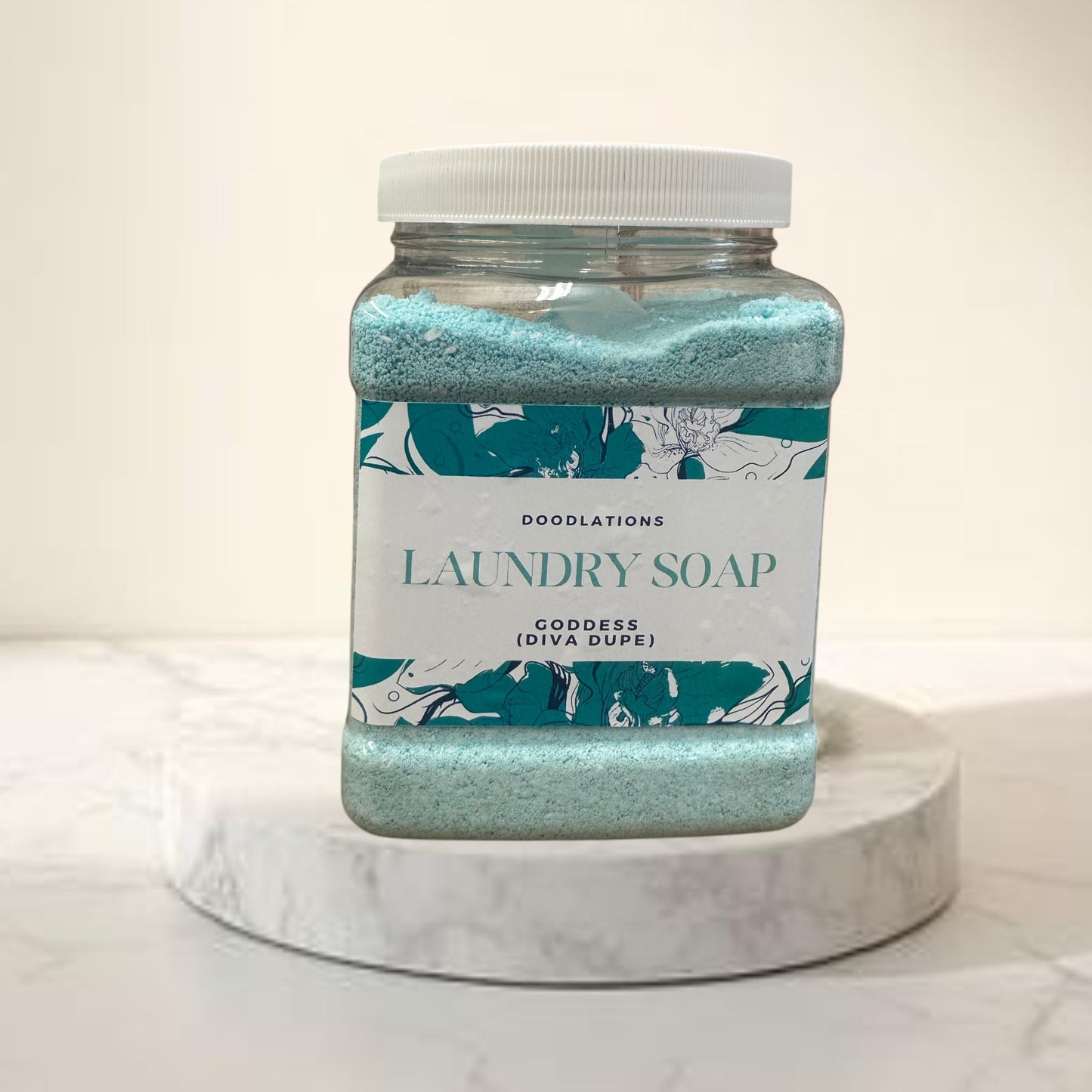 Doodlations Goddess luxury laundry powder in a clear jar with a white lid, featuring a teal floral label. The powder is light blue.