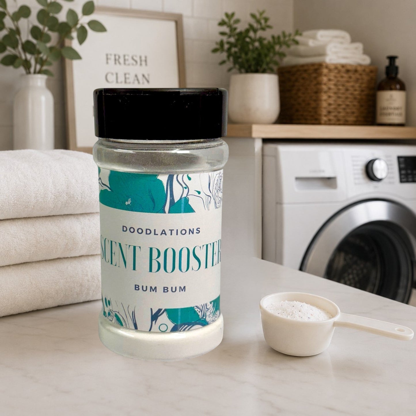 Doodlations Bum Bum laundry scent booster in a clear plastic jar with a black lid, on a marble counter next to stacked white towels.