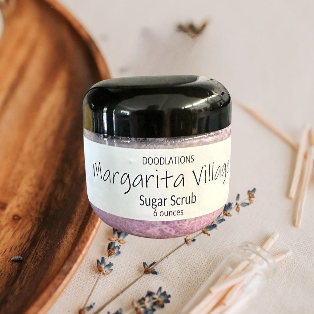 Doodlations Margarita Village sugar scrub in a clear jar with a black lid, featuring a light purple scrub and a white label.
