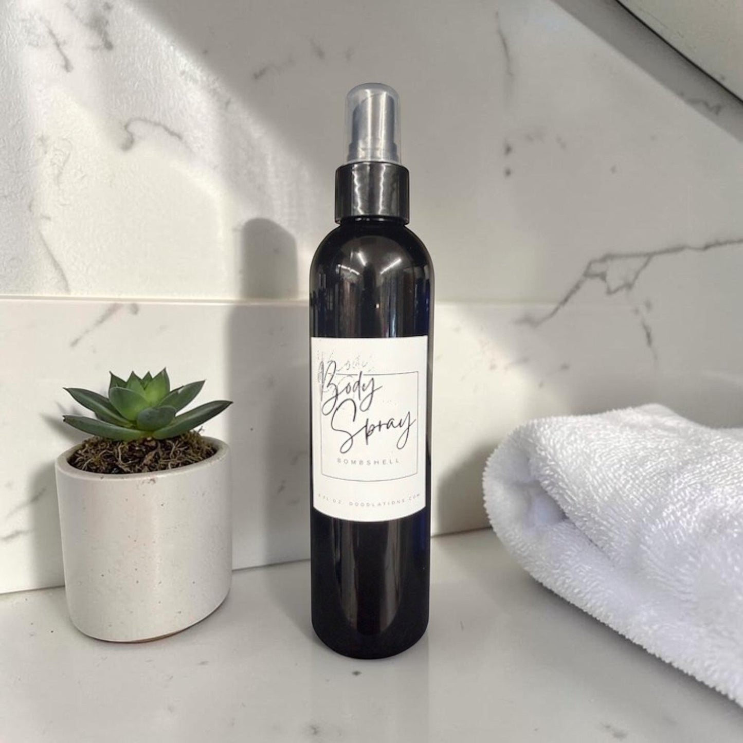 Doodlations Moisturizing Body Spray in a dark bottle with a white label, next to a small succulent and a white towel.