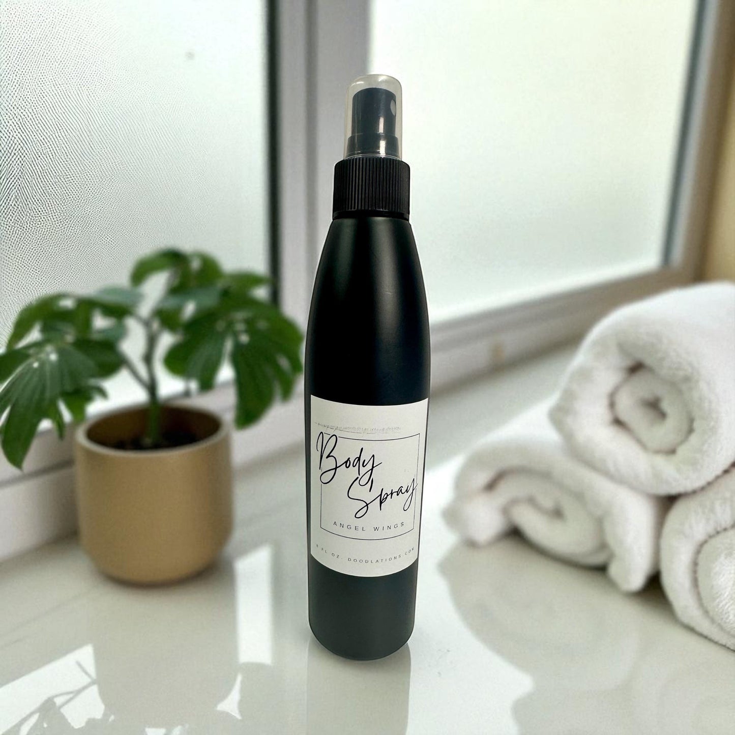 Doodlations Coffee Bar & Boutique Angel Wings moisturizing body spray in a black bottle with a clear cap, next to a plant and rolled towels.